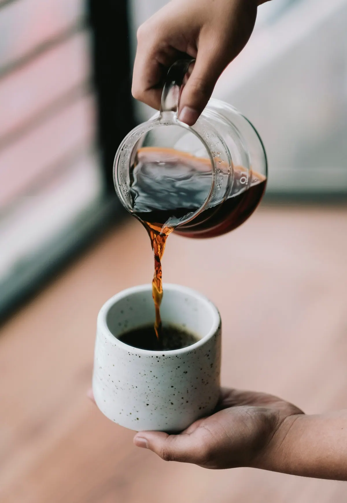 A hand pours fresh coffee from a glass server into a stone-grey cup by a window